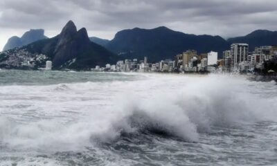 Grandes ondas quebrando na praia do Rio de Janeiro, com prédios e montanhas sob céu nublado.