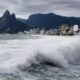 Grandes ondas quebrando na praia do Rio de Janeiro, com prédios e montanhas sob céu nublado.