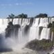 Vastas cataratas com quedas d'água brancas, rochas escuras, vegetação exuberante e céu azul.