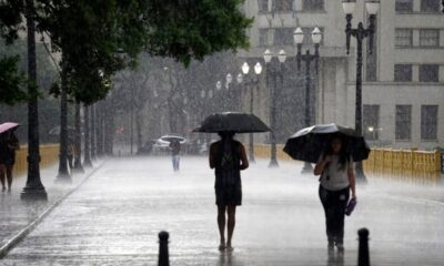 Pessoas com guarda-chuvas caminhando em rua molhada sob forte chuva em cidade.