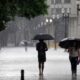 Pessoas com guarda-chuvas caminhando em rua molhada sob forte chuva em cidade.