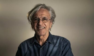 Caetano Veloso sorrindo, de óculos, com cabelo grisalho e camisa jeans.