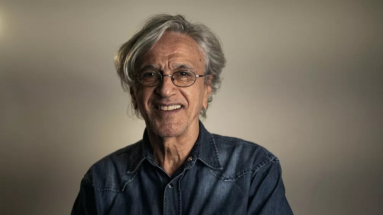 Caetano Veloso sorrindo, de óculos, com cabelo grisalho e camisa jeans.
