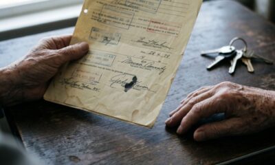 Mãos envelhecidas seguram documento antigo com assinaturas; chaves sobre mesa de madeira.
