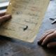 Mãos envelhecidas seguram documento antigo com assinaturas; chaves sobre mesa de madeira.