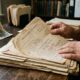 Par de mãos de um adulto folheando com cuidado uma pilha de documentos jurídicos amarelados e certidões de nascimento antigas sobre uma mesa de madeira escura e pesada