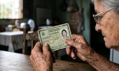 Close-up detalhado de mãos envelhecidas e calejadas segurando um documento de identidade (RG brasileiro) de modelo antigo, com o papel esverdeado apresentando marcas sutis de manuseio e tempo