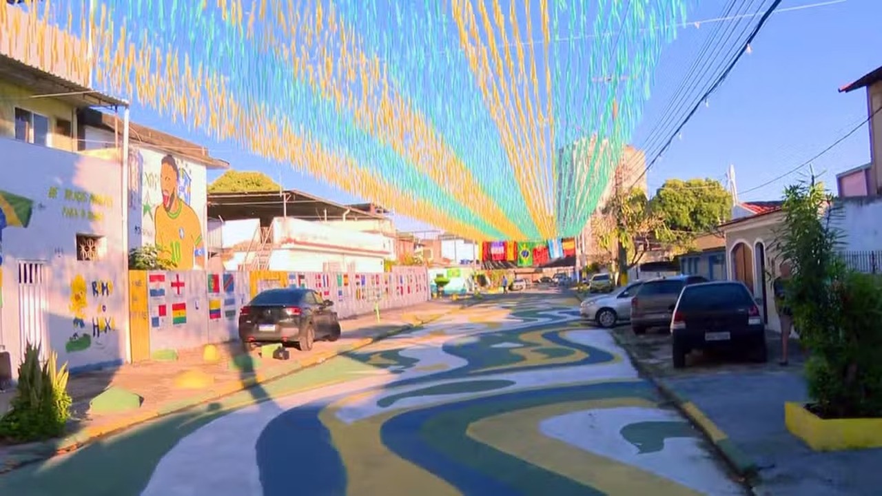 Rua decorada com fitilhos, chão pintado, mural de jogador de futebol, bandeiras de países e carros.