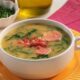 Uma tigela de caldo verde com linguiça, couve e pedaços de bacon, em um prato amarelo.