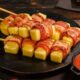 Espetos de queijo coalho envoltos em bacon dourado, servidos com molho vermelho.