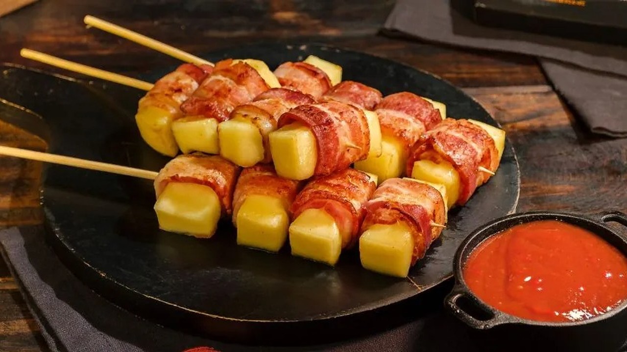 Espetos de queijo coalho envoltos em bacon dourado, servidos com molho vermelho.