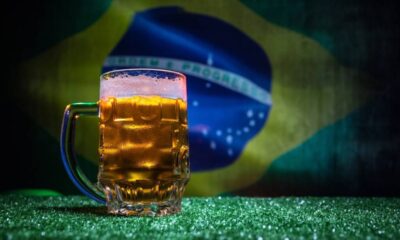 Caneca de cerveja gelada com espuma sobre grama, bandeira do Brasil desfocada no fundo.