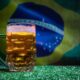 Caneca de cerveja gelada com espuma sobre grama, bandeira do Brasil desfocada no fundo.