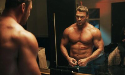 Alan Ritchson, musculoso e sem camisa, abotoa calças diante de um espelho, mulher ao fundo.