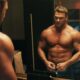 Alan Ritchson, musculoso e sem camisa, abotoa calças diante de um espelho, mulher ao fundo.