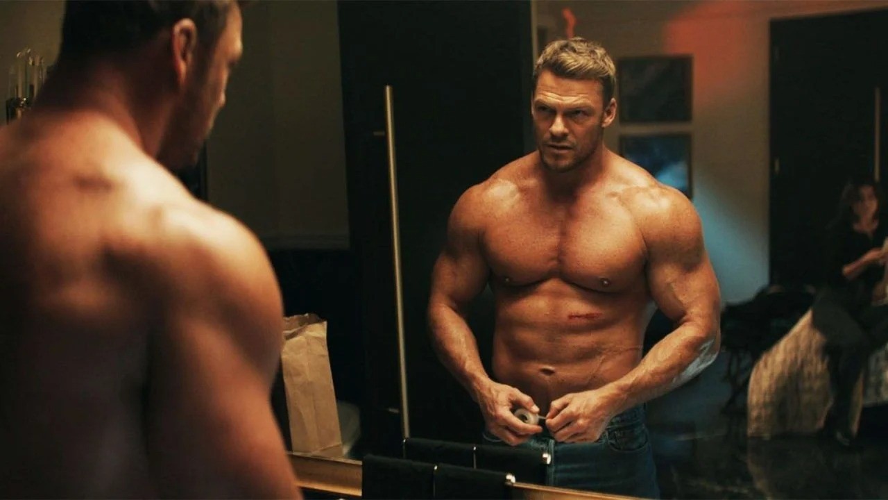 Alan Ritchson, musculoso e sem camisa, abotoa calças diante de um espelho, mulher ao fundo.