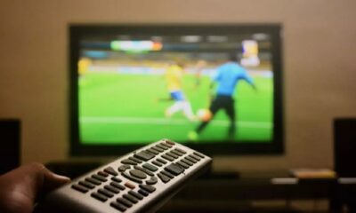 Mão segura controle remoto; TV desfocada exibe partida de futebol com jogadores e bola.