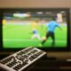 Mão segura controle remoto; TV desfocada exibe partida de futebol com jogadores e bola.