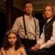 Três personagens de época, dois homens e uma mulher coroada, posam em uma sala escura e luxuosa.