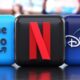 Três ícones de aplicativos: Prime Video, Netflix, Disney+. Ao fundo, TV ligada.