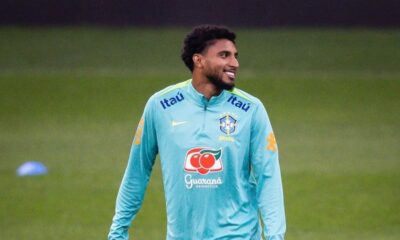 Jogador Éderson com camisa de treino da Seleção Brasileira, sorrindo em campo de futebol.