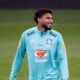 Jogador Éderson com camisa de treino da Seleção Brasileira, sorrindo em campo de futebol.