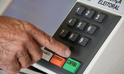 Mão pressiona botão CONFIRMA em urna eletrônica. Teclas BRANCO, CORRIGE, números com braille.