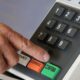 Mão pressiona botão CONFIRMA em urna eletrônica. Teclas BRANCO, CORRIGE, números com braille.