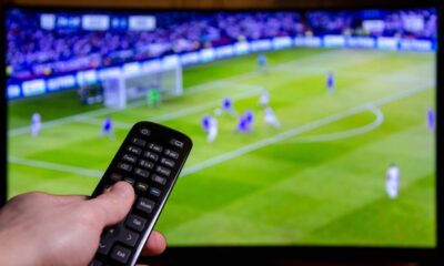 Mão segura controle remoto com botões em frente a TV exibindo jogo de futebol borrado.