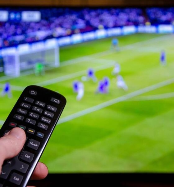 Mão segura controle remoto com botões em frente a TV exibindo jogo de futebol borrado.