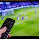 Mão segura controle remoto com botões em frente a TV exibindo jogo de futebol borrado.