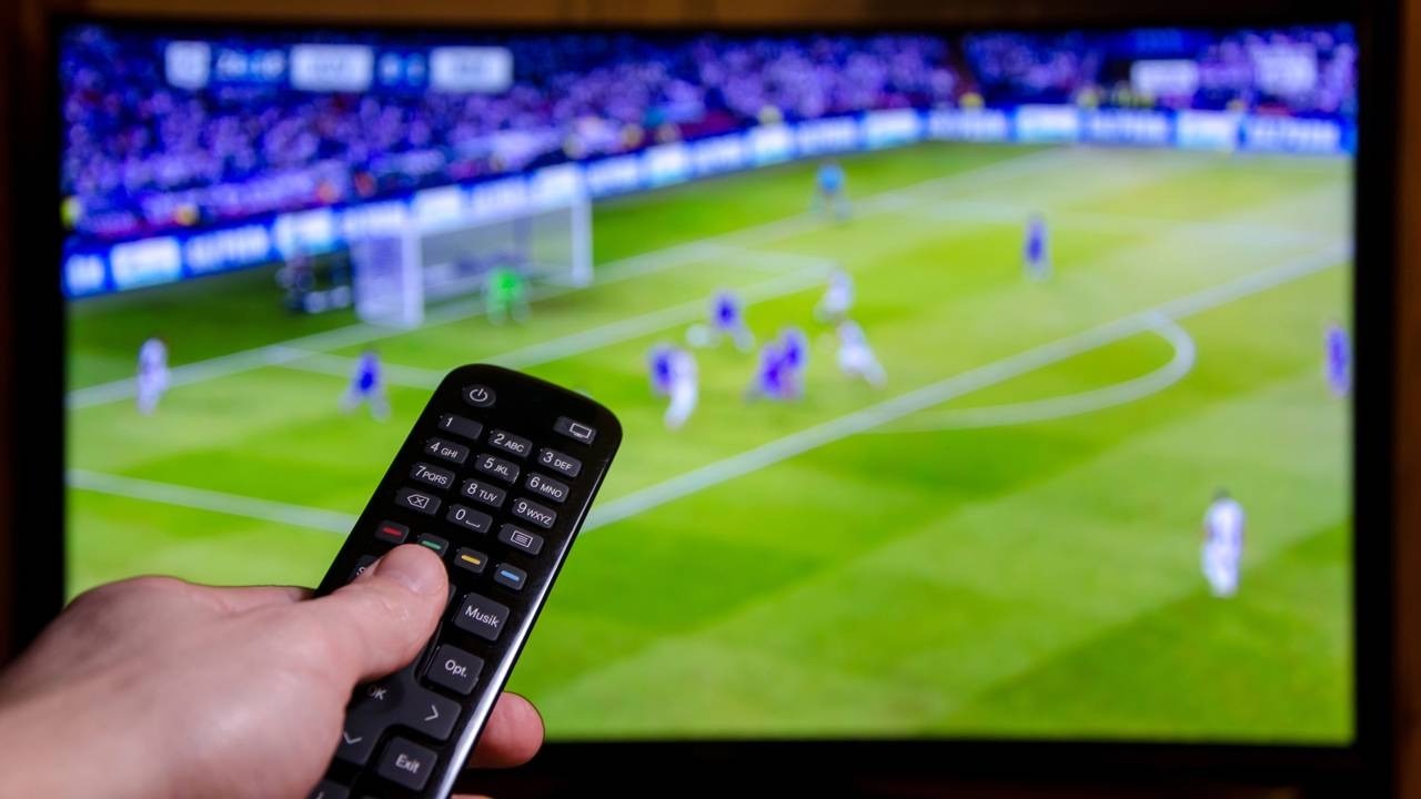 Mão segura controle remoto com botões em frente a TV exibindo jogo de futebol borrado.
