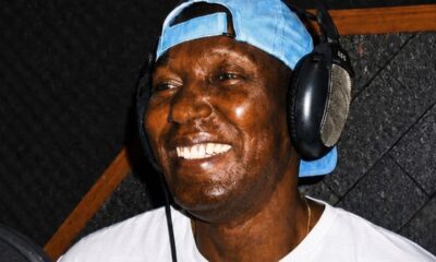 Homem negro sorridente, com fones de ouvido e boné azul, em estúdio de gravação.