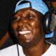 Homem negro sorridente, com fones de ouvido e boné azul, em estúdio de gravação.