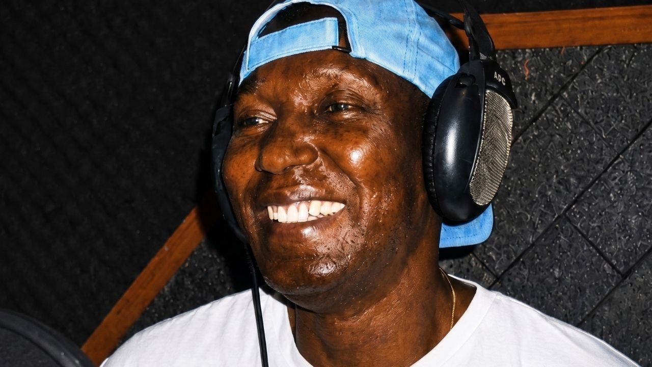 Homem negro sorridente, com fones de ouvido e boné azul, em estúdio de gravação.
