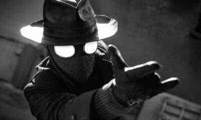 Close-up de Spider-Noir em preto e branco, com chapéu, óculos brancos, casaco e mão estendida.