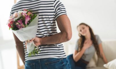 Homem esconde buquê de flores rosa e brancas atrás das costas, surpreendendo mulher sorrindo no sofá.