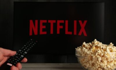 Tela de TV preta com logo NETFLIX, mão com controle remoto e tigela de pipoca.