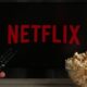 Tela de TV preta com logo NETFLIX, mão com controle remoto e tigela de pipoca.
