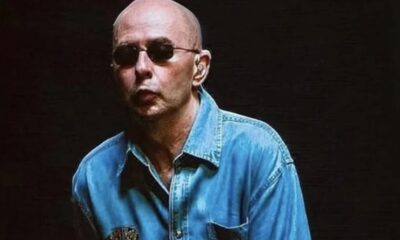 Homem careca, de óculos escuros, e camisa jeans azul em fundo escuro.