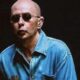 Homem careca, de óculos escuros, e camisa jeans azul em fundo escuro.