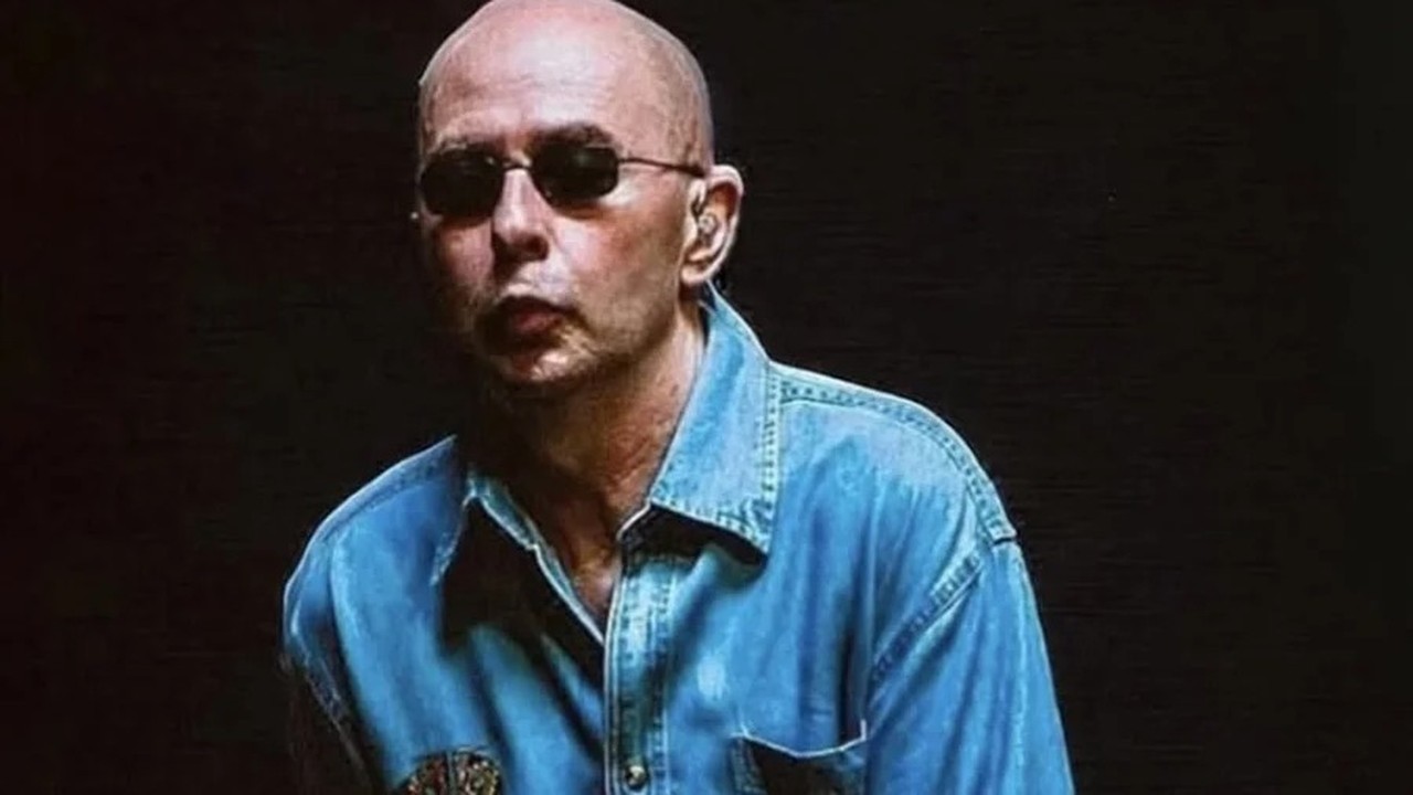 Homem careca, de óculos escuros, e camisa jeans azul em fundo escuro.