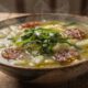Uma tigela rústica de caldo fumegante com linguiça, batata e cheiro-verde em mesa de madeira.