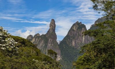 Montanhas rochosas com destaque para o pico Dedo de Deus e vegetação densa sob céu azul.
