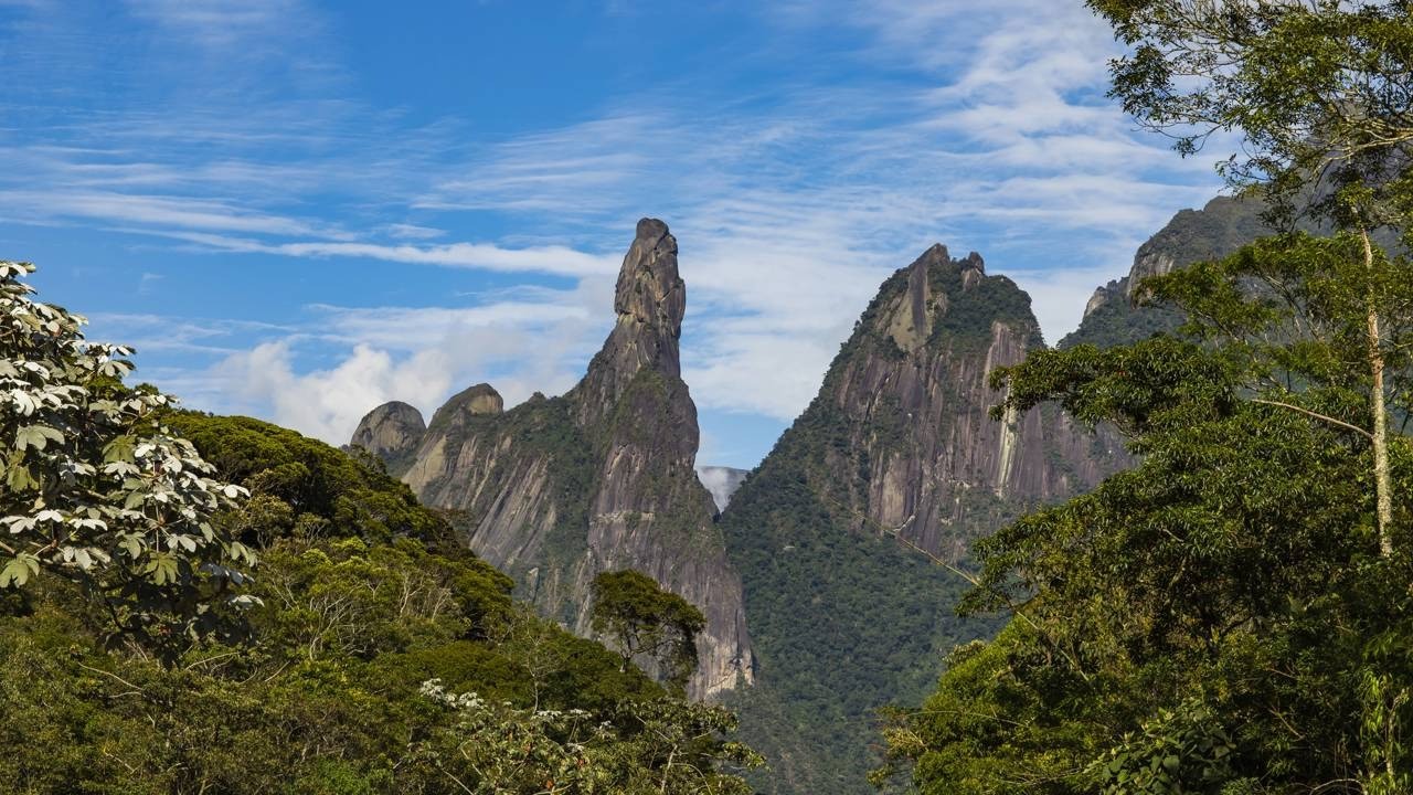 Montanhas rochosas com destaque para o pico Dedo de Deus e vegetação densa sob céu azul.