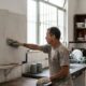 Homem com luvas aplica massa em parede cinza da cozinha, com azulejos antigos na bancada.