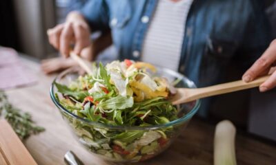 Mãos misturam salada fresca e colorida em uma tigela de vidro com colheres de madeira.