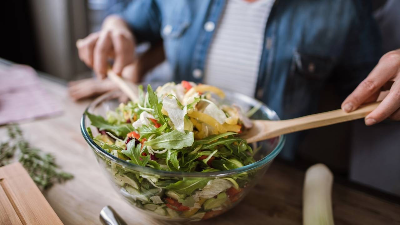 Mãos misturam salada fresca e colorida em uma tigela de vidro com colheres de madeira.
