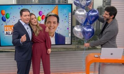 Estúdio de TV. Alan Severiano, Jacqueline Brazil e colega com balões, festa em telão.
