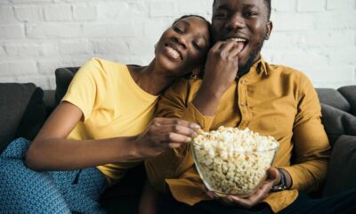 Casal negro feliz no sofá, comendo pipoca de uma tigela de vidro.